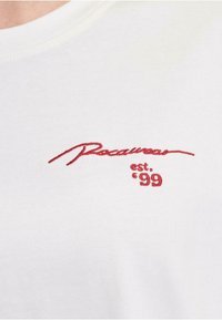 White cotton t-shirt featuring red embroidered text "Rocawear" and "est. '99" in a cursive style, smooth texture, and classic crew neckline.