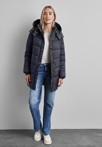 Abrigo de plumas marinero con capucha y cuello negro con estampado, con cierres a presión y bolsillos laterales, llevado sobre jeans de mezclilla claros y zapatillas.