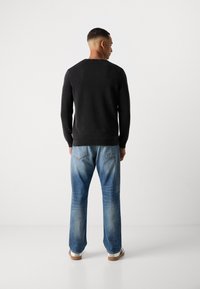 Schwarzer gestrickter Pullover mit rundem Ausschnitt, kombiniert mit hellblauen Jeans mit einem ausgewaschenen Finish und genähten Gesäßtaschen. Weiße Schuhe mit brauner Sohle.