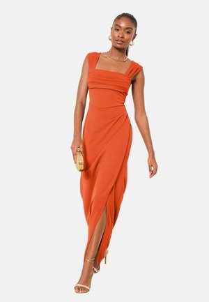 Žena v oranžových dlhých šatách bez rukávov s predným rozparkom, drží zlaté psaníčko a nosí zlaté sandále na vysokom opätku a šperky.