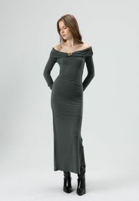 Robe vert foncé à manches longues et épaules dénudées, ornée de fronces et d'un accent doré au niveau de l'encolure, associée à des bottes à talons noires.