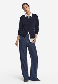 Marineblauer Strickjacke mit Knöpfen, über einem weißen Hemd getragen, kombiniert mit hoch taillierten dunklen Jeans aus Denim. Glatte Textur, klassisches Design.