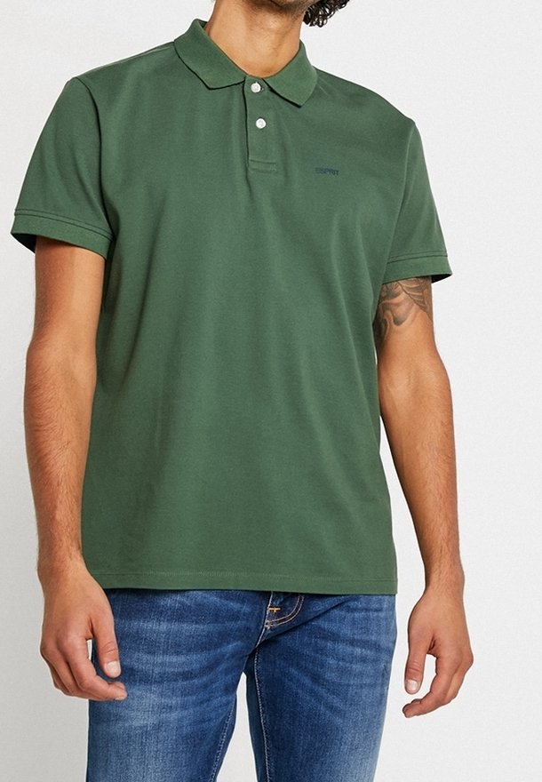 Grön bomullspiké med krage, treknappsspäning och broderad logotyp. Kombinerad med blå denimjeans.