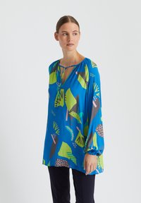 Blouse bleue avec des motifs abstraits audacieux en vert, rose et noir. Dispose d'un décolleté en V et de longues manches froncées. Tissu léger.
