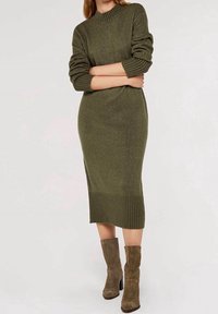 Femme portant une robe midi en maille vert olive avec poignets et ourlet côtelés, associée à des bottines en daim marron assorties.