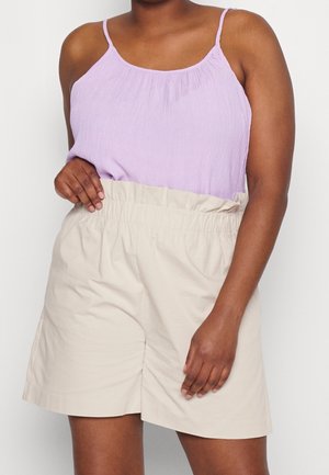 Kobieta ubrana w liliowy bezrękawnik i beżowe szorty z wysokim stanem i elastycznym pasem, stojąca na jednolitym tle.