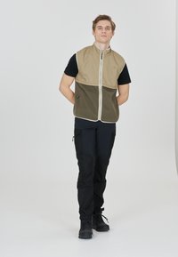 Beige och mörkgrön zippered väst med ärmlös design, fleece-nedre del och sidofickor, bärs över en svart t-shirt och svarta byxor.