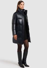 Padded crni parka s sjajnom završnom obradom, visokim ovratnikom i zip džepovima, nosi se preko crne pletene haljine i uparen je s crnim čizmama do koljena.
