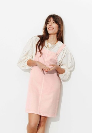 Femme souriante, portant un chemisier blanc à manches bouffantes et une robe salopette rose sans manches, avec un collier à chaîne dorée épais.