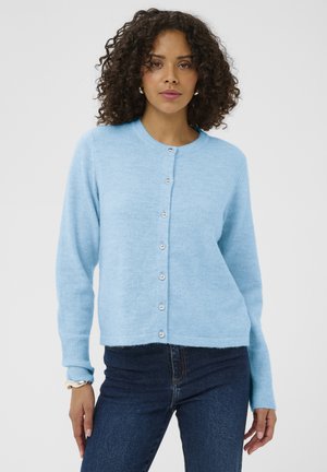 Kvinde med krøllet hår iført en lyseblå knapåbnet cardigan og mørkeblå jeans foran en ensfarvet hvid baggrund.