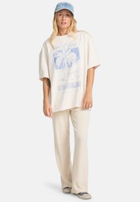 Lysbeige oversized T-shirt med blå grafisk tekst og palme-design, parret med cremefarvede ribbede bukser med vide ben og grå sneakers.