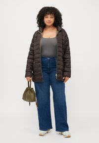 Brown puffer jacket with quilted design, zip closure, paired with high-waisted blue jeans and a green drawstring bag.