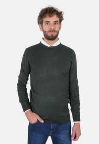 Maglione verde scuro lavorato a maglia con scollo rotondo e polsini a costine, abbinato a una camicia bianca con colletto sotto. Jeans di jeans blu.