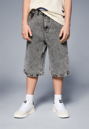 Person iført syrevasket grå denimshorts, hvide crew sokker og hvide sneakers med sorte detaljer, står mod en ensfarvet grå baggrund.