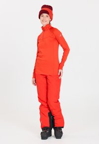 Rød langærmet zip-up top og matchende bukser, begge i et glat, teknisk stof. Sorte skistøvler med spænder fuldender outfittet.