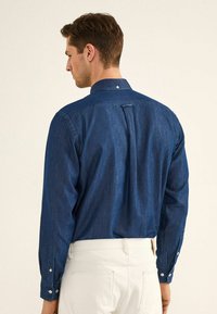 Camicia di jeans blu con maniche lunghe, polsini con bottoni e una piega sul retro. Presenta un colletto classico e un singolo bottone nella parte posteriore.