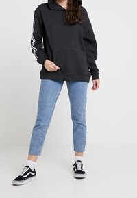 Sudadera negra con un bolsillo frontal y capucha con cordón, combinada con unos vaqueros recortados de color azul claro y zapatillas negras con detalles blancos.