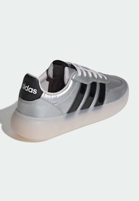 Silverfärgad syntetisk sneaker med svarta läderdetaljer, utrustad med tre svarta ränder, vit sula och vita skosnören. Logotyp synlig på hälen.