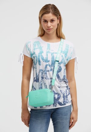 Junge Frau trägt ein weißes T-Shirt mit Grafik und blaue Jeans, sie hat eine mintgrüne Umhängetasche mit verstellbarem Riemen dabei.