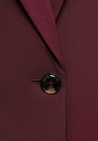 Burgundský látkový blazer s texturovaným povrchem, s klopami a lesklým černým knoflíkem se čtyřmi dírami.