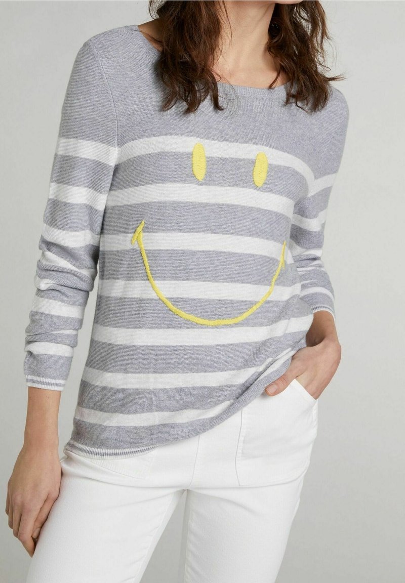 Femme portant une chemise à manches longues rayée grise et blanche avec une broderie visage souriant jaune, associée à un pantalon blanc, main dans la poche.