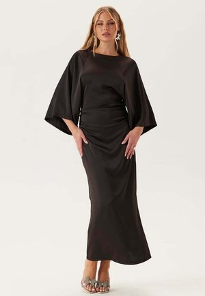 Kvinde iført en lang sort kjole med vide ærmer, stående med hænderne nær hofterne, accessoriseret med øreringe og sølvfarvede åbne tåhæle.