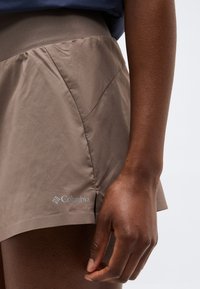 Κοντινό πλάνο ενός χεριού που ακουμπά κοντά σε χακί σορτς Columbia με φαρδιά ζώνη μέσης, πάνω σε απλό φόντο.