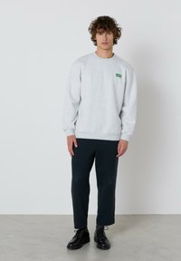 Osoba s kučeravými vlasmi, oblečená v svetlo šedej mikine s zeleným textom, čiernych krátkych nohaviciach a čiernych šnurovacích topánkach, stojí na šedom podlaží.