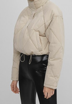 Giacca trapuntata beige con colletto alto, tasca frontale e orlo con cordino. Abbinata a leggings neri lucidi.
