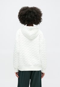 Felpa bianca trapuntata con un motivo a onde testurizzato, dotata di un ampio cappuccio e polsini a costine. Il modello indossa pantaloni larghi di un verde scuro.