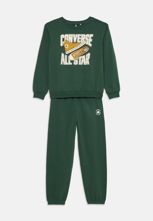 Zaļš sporta krekls un bikses ar dzelteno Converse apavu grafiku un tekstu "CONVERSE ALL STAR". Ribotas aproces un jostasvieta.