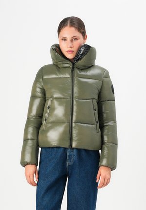 Vrouw in een olijfgroene glanzende pufferjack met hoge kraag en blauwe jeans, staand tegen een effen witte achtergrond.