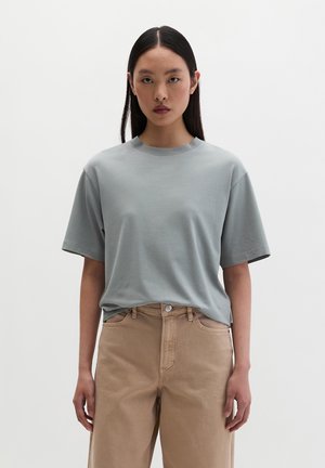 Kobieta z długimi czarnymi włosami, ubrana w luźny jasnoszary t-shirt wsunięty w beżowe spodnie z wysokim stanem, stojąca na jednolitym białym tle.