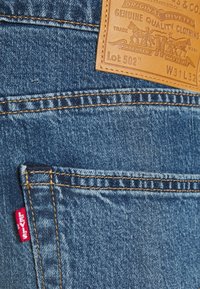 Los jeans de mezclilla azul de Levi's presentan un parche de cuero, costuras naranjas y una pequeña etiqueta roja de Levi's cerca del bolsillo. Etiquetados como Lot 502, W31 L32.