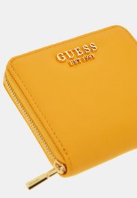 Kollane tõmblukuga rahakott, mis on valmistatud siledast sünteetilisest materjalist, esiküljel on kuldne reljeefne logo tekstiga "GUESS EST 1981".