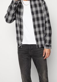 Sort og hvidternet flannel-skjorte, knappet, lagdelt over en hvid T-shirt, kombineret med mørke jeans. Bemærkelsesværdige knapdetaljer og krave.