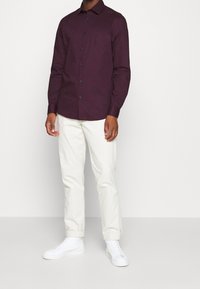 Donkerpaars overhemd met lange mouwen, puntige kraag en zwarte knopen, gecombineerd met lichtgekleurde broek en witte sneakers.
