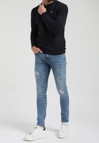 Pull à manches longues noir texturé, associé à un jean slim délavé bleu clair avec des effets usés, et des baskets blanches. Détail simple du logo sur le pull.