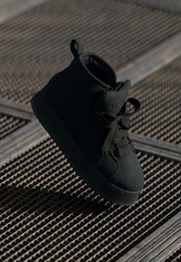 Svart høyhælte sneakers laget av teksturert semsket skinn med rund tå, snøring og en løkke ved kragen. Flat gummisåle.