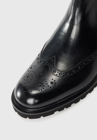 Bota de cuero negra con un acabado pulido. Presenta perforaciones decorativas y un corte superior en forma de ola. Suela de goma gruesa para mayor durabilidad.