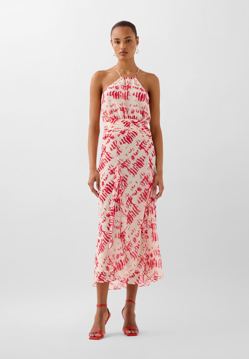 Maxi-jurk van lichtcrème stof met een rood abstract patroon. Beschikt over verstelbare schouderbanden en een vloeiende silhouet, gecombineerd met rode gehakte sandalen.