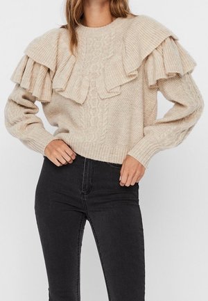 Beige kabelgebreide trui met grote gelaagde ruches aan de hals, gecombineerd met hooggetailleerde zwarte skinny jeans.