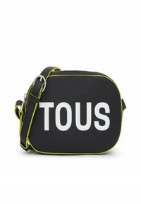 Bolso cruzado de cuero negro con el texto en relieve "TOUS" en blanco. Presenta acentos en verde lima y una correa desmontable. Acabado de textura suave.