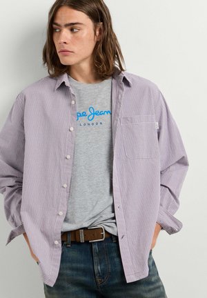 Jong persoon met schouderlang haar draagt een grijs grafisch T-shirt onder een opengeknoopt klein geruit overhemd met jeans en een bruine riem.