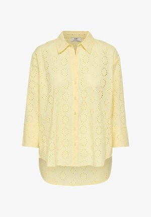 Camisa amarilla clara con botones y cuello, con patrones florales recortados y mangas de tres cuartos de longitud; dobladillo curvado.