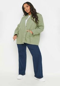 Olivgrön, knapp-up jacka med två framfickor, stylad över en grå topp; matchad med mörkblå vida jeans och vita skor.