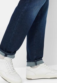 Donkerblauwe denim jeans, opgerold bij de enkels, gecombineerd met witte sneakers met perforaties en veters, tegen een lichte achtergrond.