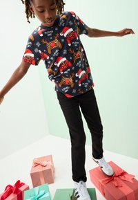 Schwarzes grafisches T-Shirt mit bunten Gaming-Motiven, darunter Controller und Rentiere, kombiniert mit schwarzer Hose und weißen Sportschuhen.