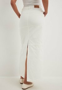 Jupe maxi en denim blanc avec une taille ajustée, une fente latérale marquée et des détails cousus. Texture lisse et patch logo beige à l'arrière.