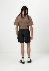 Brun kortærmet t-shirt, sorte shorts med snøre og sorte sneakers med hvide striber. Positioneret med en neutral baggrund.